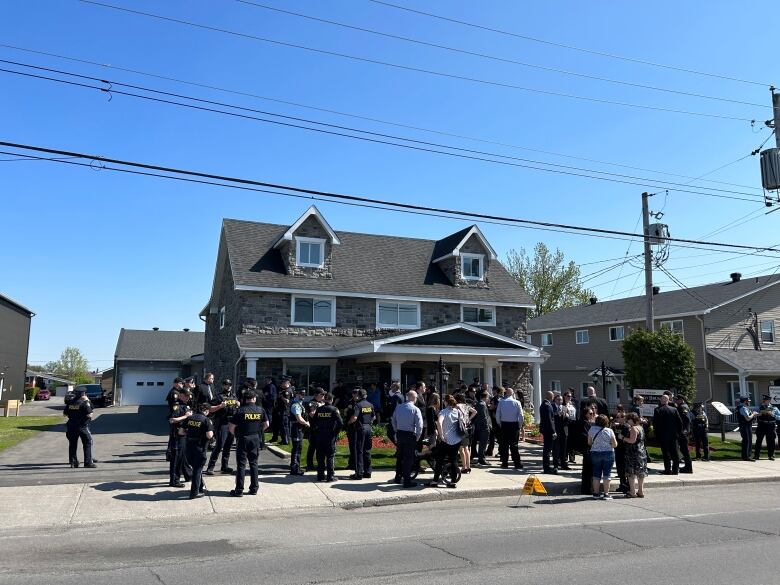 Groups of police officers stand on the sidewalk and street outside a home. 