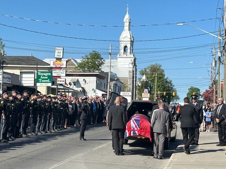 People walk behind a hearse on a busy city street. A casket draped with a red flag can be seen inside the hearse, and dozens of police officers stand in a line on the sidewalk.