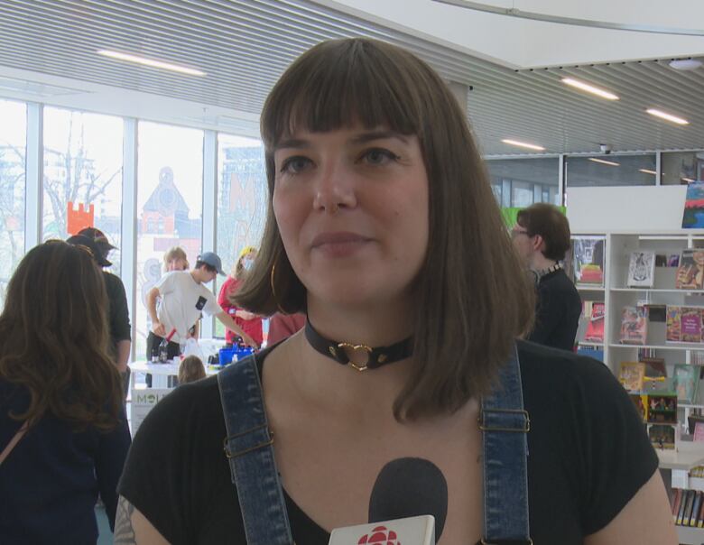 A woman with bangs who is wearing overalls speaks to a reporter. 