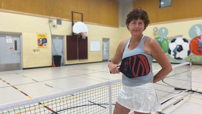 Woman with short hair and tennis attire stands in front of net with paddle across her chest, smiling. 