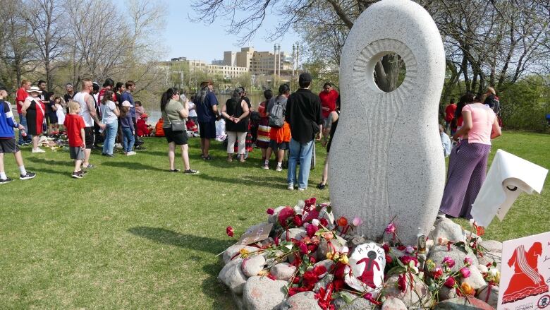 A memorial statue is pictured, covered with flowers laid by marchers, who stand facing away from it, toward a river.