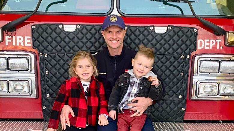 A man sits on the front bumper of a fire engine, with two children on his lap. They are all looking at the camera and smiling.