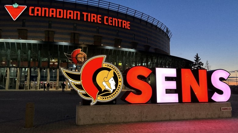 A red and white lit-up SENS sign in front of a hockey arena.