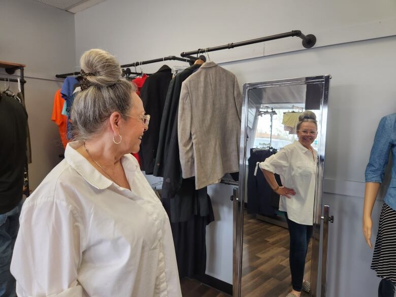 A woman in a white shirt stands in front of a mirror with her hand on her hip.