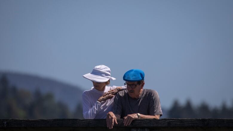 Two older adults, a man wearing a blue cap and a woman wearing a white sun hat, stand outside in Vancouver on a sunny day.