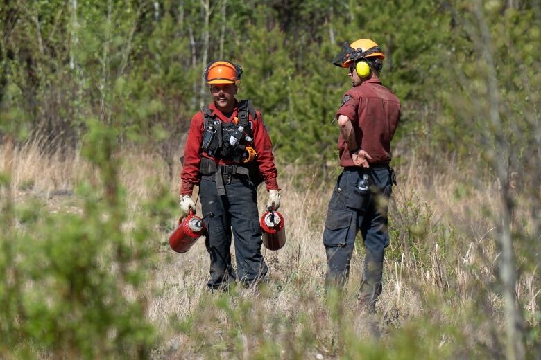 Two firefighters stand in a grassy area and hold equipment.