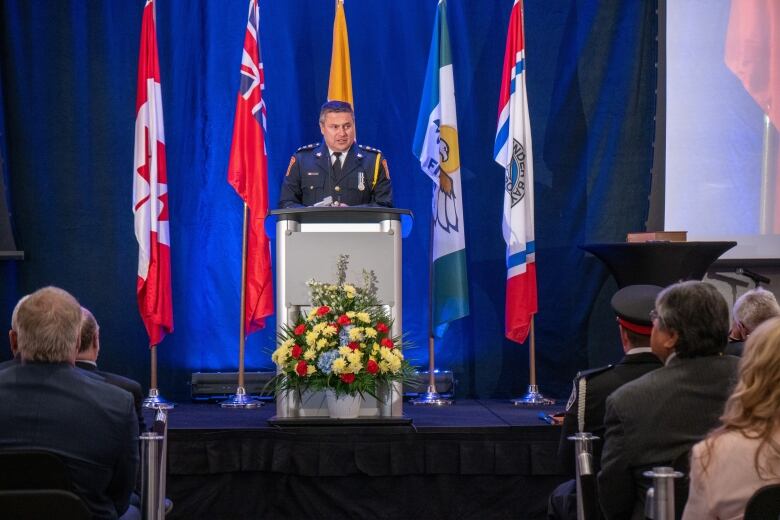 A man in a formal police uniform speaks at a podium while a crowd listens.