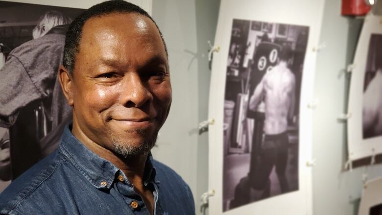 A man wearing a blue shirt smiles into the camera with a gallery of black and white photos hang in the background.