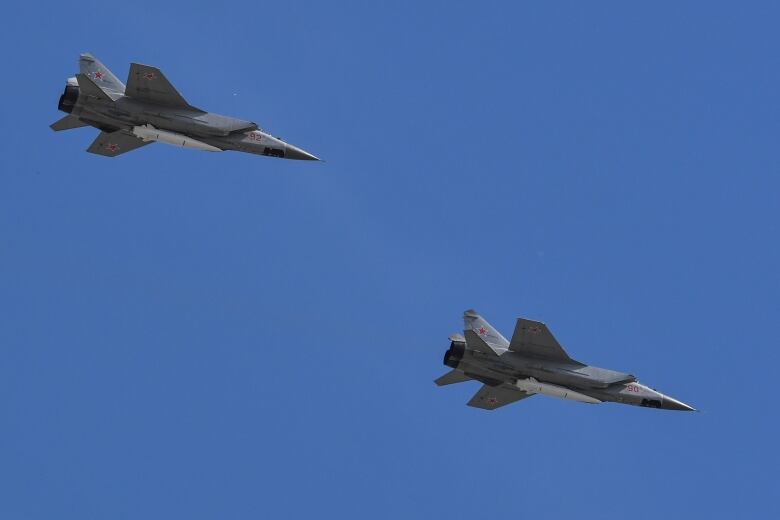 Two grey fighter jets, with a large missile attached on the bottom of each, flying in the blue sky.