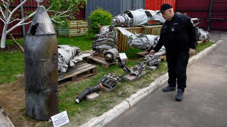 A man in a a black jacket and black pants inspects large pieces of metal debris and a large missile head standing on the grass. 