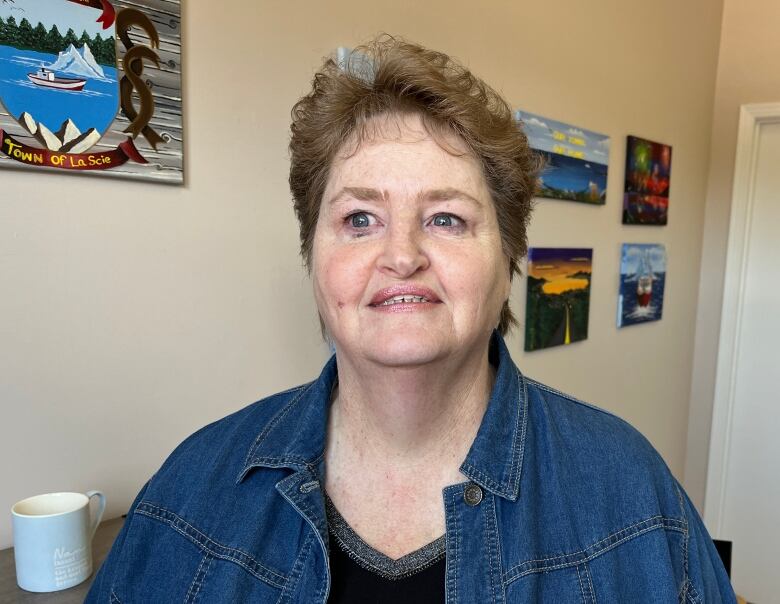 A woman in a black shirt and jean jacket stands in front of a wall with painted landscapes.