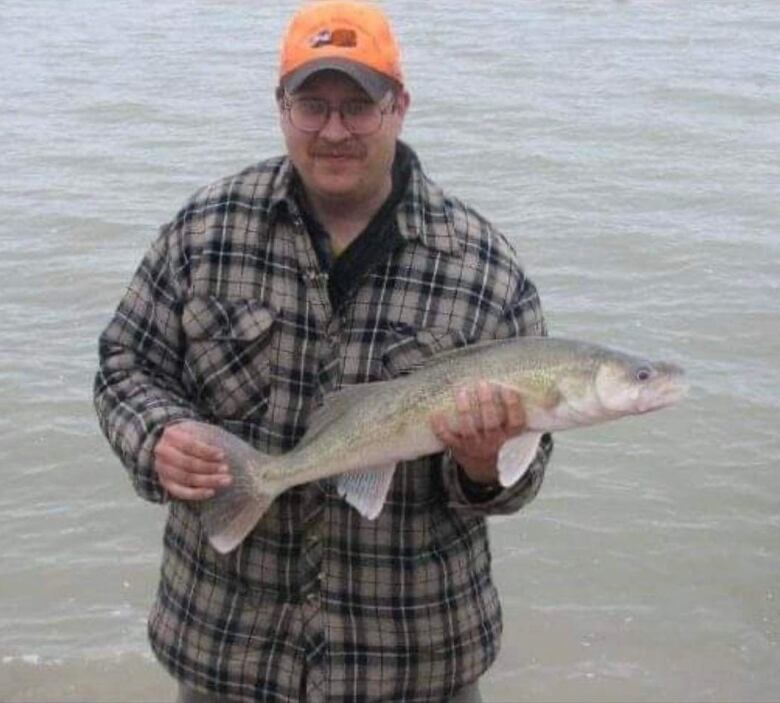 A man in a grey flannel sweater, glasses and an orange ball cap holds a fish while standing near a lake.