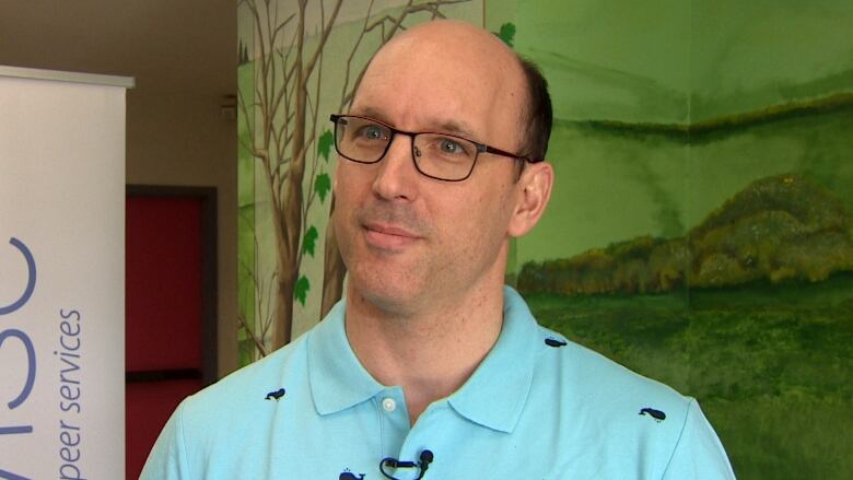 A man wearing a blue collared shirt standing in front of a green backdrop. 