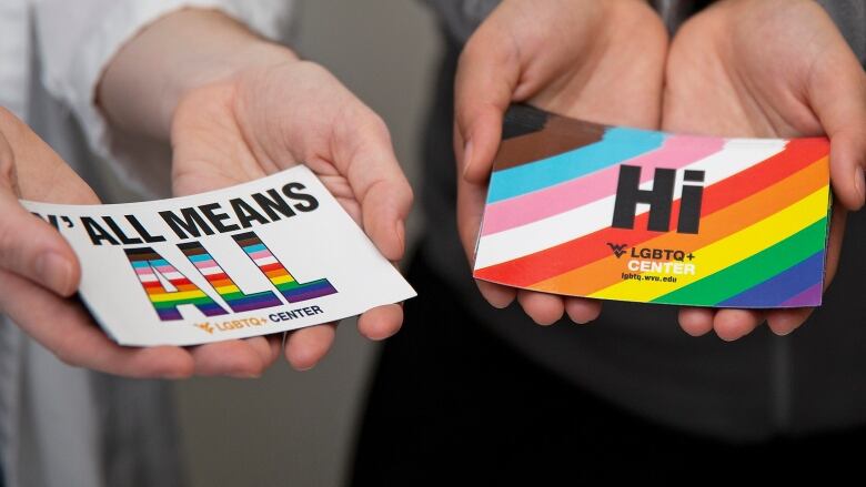 Three hands hold stickers and pins with pride flags and LGBTQ+ slogans on them.