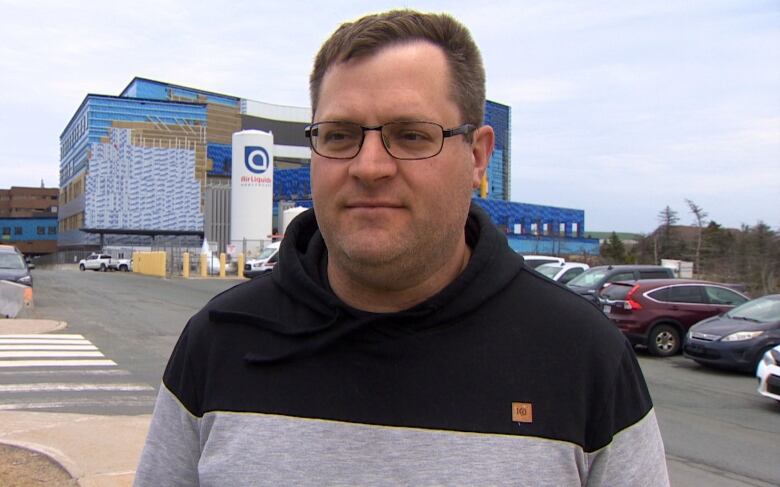 A man wearing glasses and a black and grey sweater standing near a parking lot. In the background is a building under construction. 