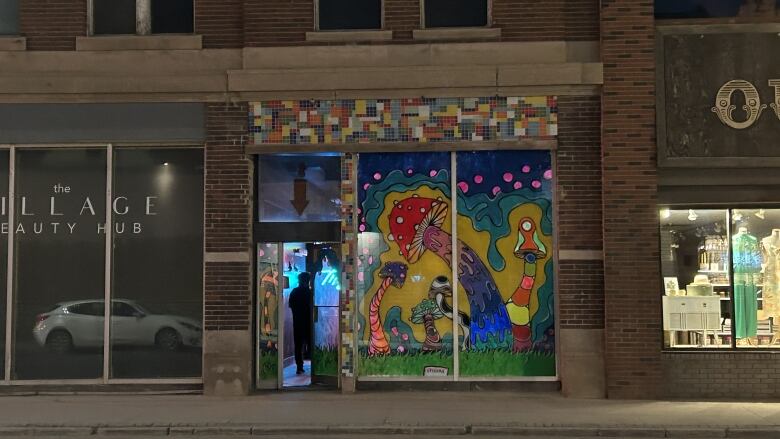 A storefront with coulourful mushroom artwork in its window is shown on a commercial street at night.