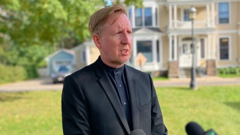 A man with blond hair in a dark suit stands outside talking to reporters.