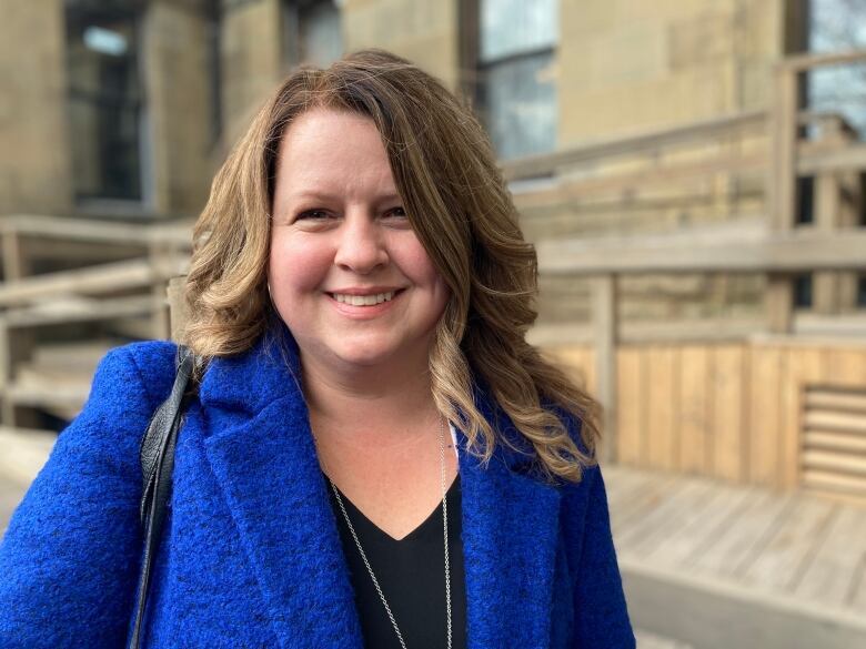 A woman with long light-brown hair, in a blue suit and black shirt stands outside the legislature and smiles for the camera.