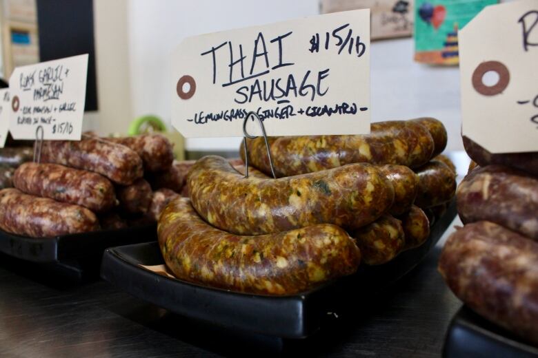 Stacks of sausages sit on three plates, each with a different sign. The central one in focus reads 'Thai sausage  lemongrass, ginger + cilantro.' It costs 15 dollars per pound.