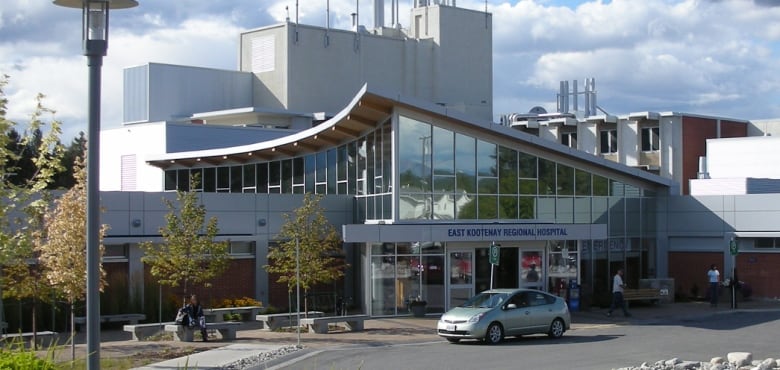 A silver car is parked outside the front doors of a grey hospital on a sunny day. A person is visible sitting at a picnic table.