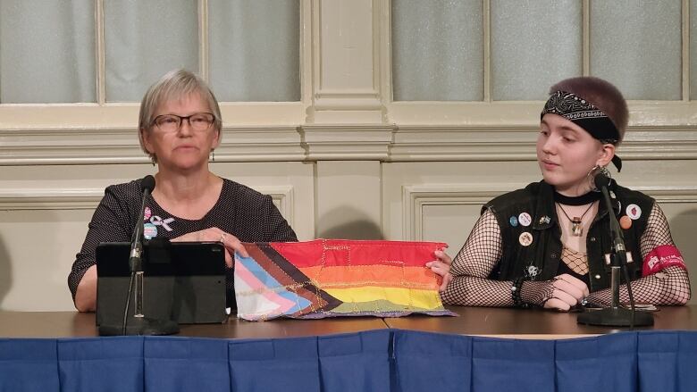 At a press conference, a woman with grey hair, left, holds up a Pride flag which had been ripped up. To her right, a high school student looks on.