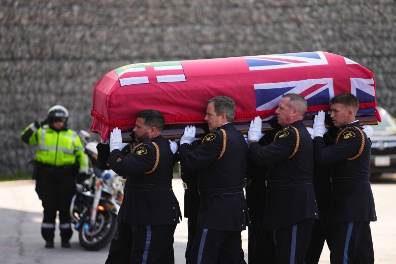 A group of police officers carry a casket wrapped in a red Ontario flag. Another officer salutes in the background next to a motorcycle. 