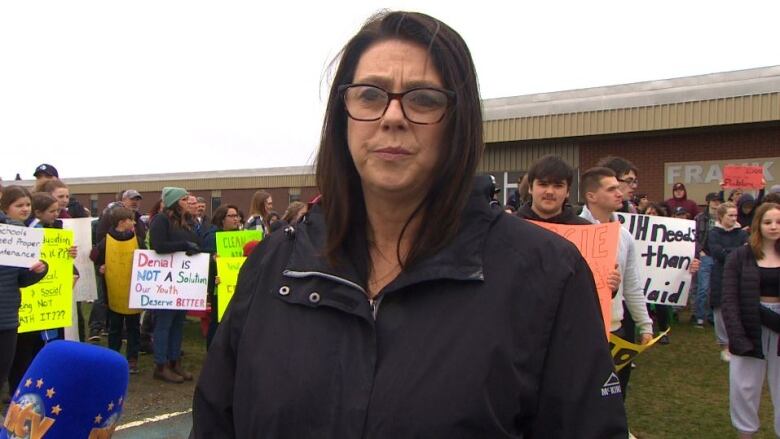 A woman wearing glasses and a black jacket stands in front of a crowd of people holding protest signs. 