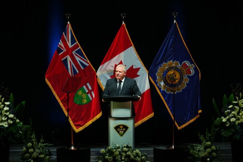 A politician speaks at a funeral in front of a black drape and 3 flags.