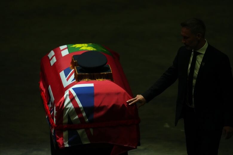 A man in a suit touches a casket wrapped in a red Ontario flag.