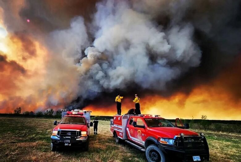 Firefighters standing on the back of red firetrucks look back at a sky full of flames and smoke in this surreal wide-angle photo.