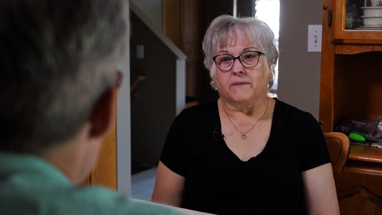 A woman with grey hair wearing a black shirt and glasses sits at her kitchen table.