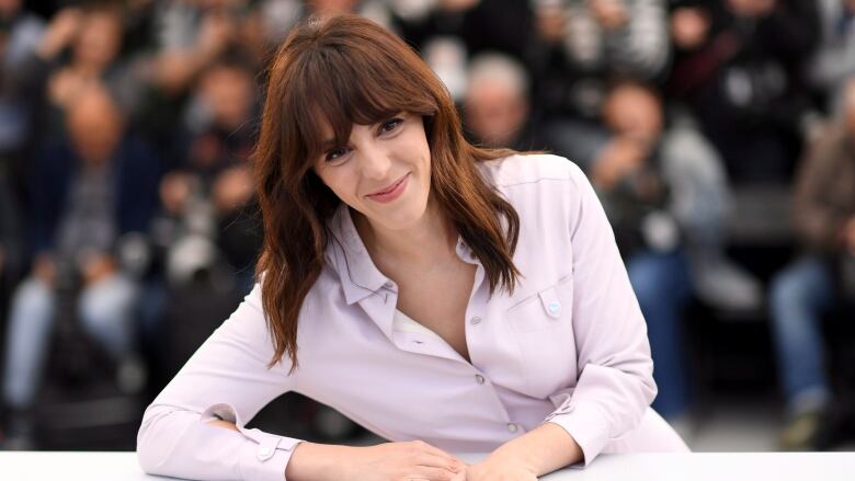 A woman with red hair wearing a light button down leans onto a table.