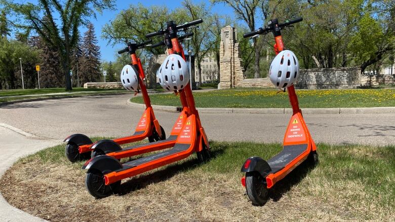 Four bright orange e-scooters are sitting on the grass with helmets hanging from the handle bars.