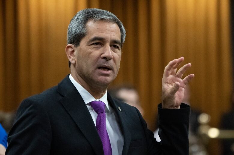 A man in a suit and a purple tie stands up and gestures.