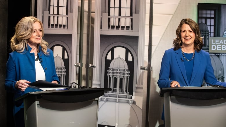 Two women wearing blue suits stand at podiums opposite each other.