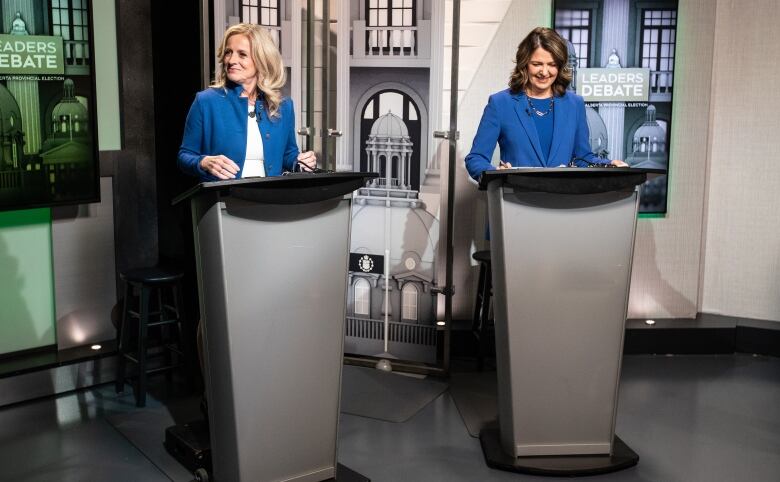 Rachel Notley looks off and to the right, while Danielle Smith checks her notes, both standing behind lecterns.