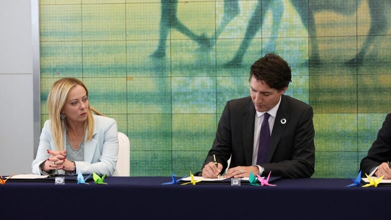 A man in a dark suit sits at a table, signing a document with a pen. To his right, a woman in a pale blue suit, clasps her hands.