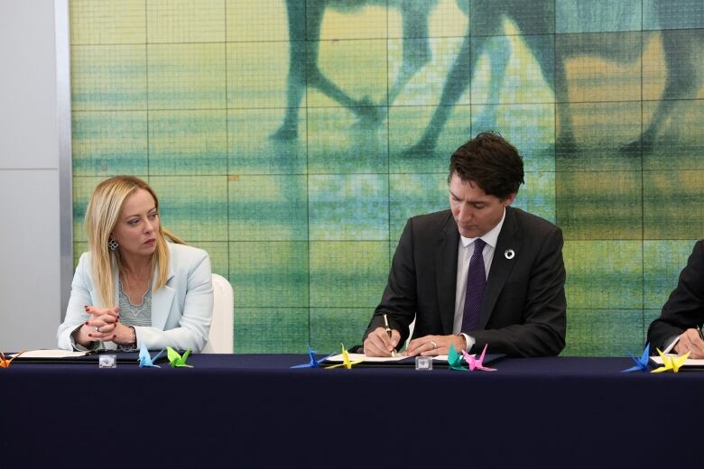 A man in a dark suit sits at a table, signing a document with a pen. To his right, a woman in a pale blue suit, clasps her hands.