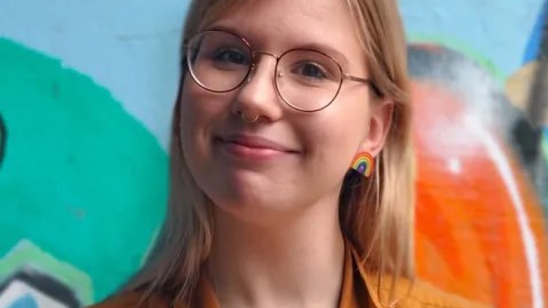 A person with blond hair and wire-framed glasses is smiling. She is wearing a rainbow earring.