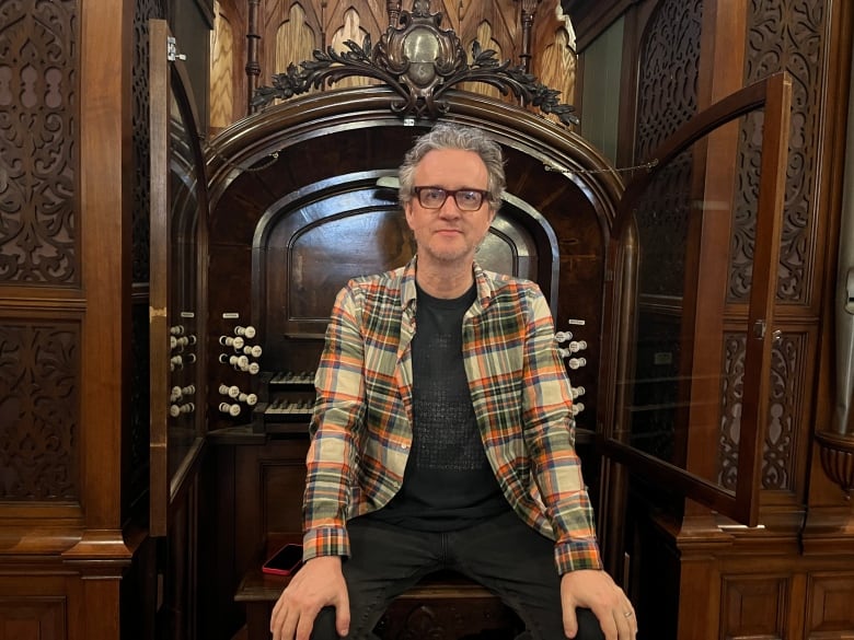 A man wearing a plaid shirt and t-shirt sits on a bench in front of an antique pipe organ. 