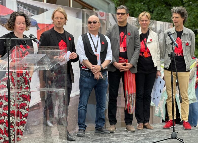 A woman speaks at a podium while four men and a woman look on.