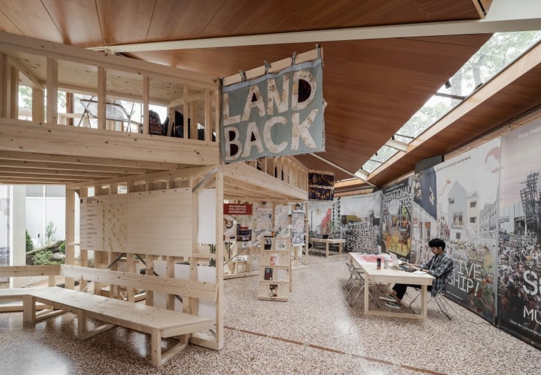 A person sits at a table inside a pavilion surrounded by posters. A sign that reads 'Land Back' hangs from a wooden beam.