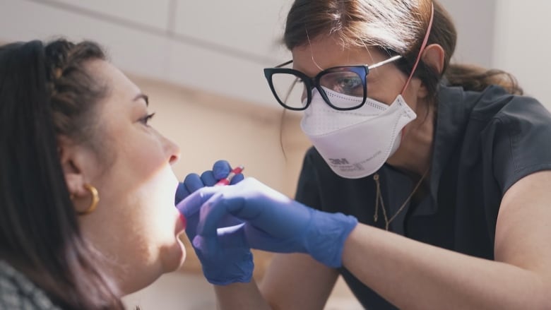 On the left, a white woman with long dark hair and gold hoop earrings opens her mouth as a masked female dentist with dark framed glasses examines her teeth from the right.