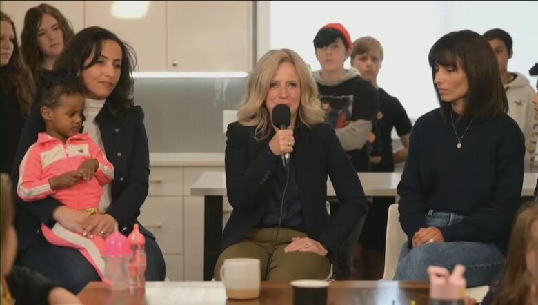 A blonde woman in a black blazer speaks into a microphone surrounded by sitting and standing children, teens and other women.