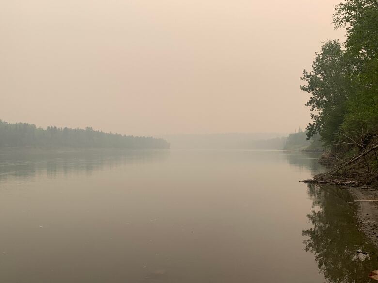 A hazy, smoky sky over a river with green trees on the banks.