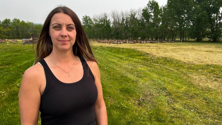 A woman with brown hair and a sleeveless black shirt looks at the camera, with a field of grass behind her.