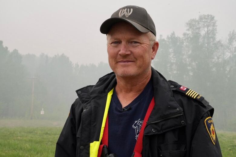 A man in a black jacket and a brown baseball cap stands outdoors with a slight smile on his face. Behind him, smoke hangs in the air.