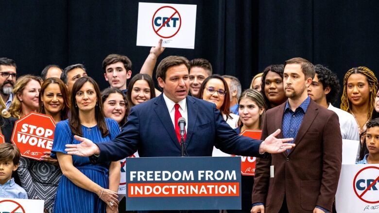 A man speaks at a podium with text reading 'Freedom from indoctrination' as people stand around him holding signs that read 'Stop woke' or show the acronym 'CRT' with a line through it.