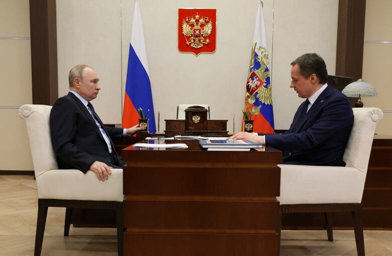 Two people are seen seated at a table, as Russian flags are seen next to them.