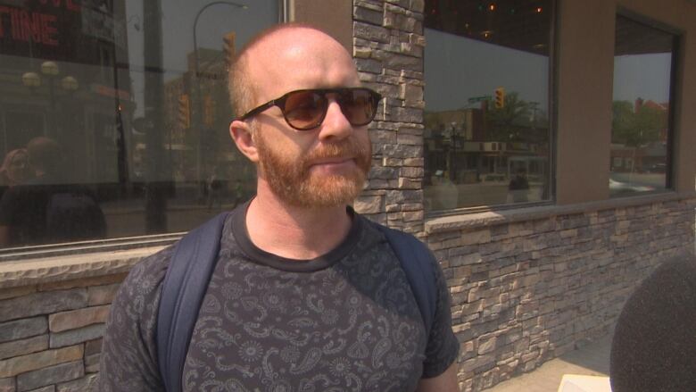 A man with short hair and a beard speaks with a reporter on a street side.